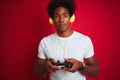 Afro gamer man playing video game using joystick headphones over isolated red background with a confident expression on smart face Royalty Free Stock Photo