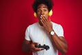 Afro gamer man playing video game using joystick headphones over isolated red background cover mouth with hand shocked with shame Royalty Free Stock Photo