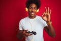 Afro gamer man playing video game using joystick headphones over isolated red background doing ok sign with fingers, excellent Royalty Free Stock Photo