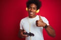 Afro gamer man playing video game using joystick headphones over isolated red background happy with big smile doing ok sign, thumb Royalty Free Stock Photo