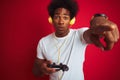 Afro gamer man playing video game using joystick headphones over isolated red background pointing with finger to the camera and to Royalty Free Stock Photo