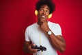 Afro gamer man playing video game using joystick headphones over isolated red background serious face thinking about question, Royalty Free Stock Photo