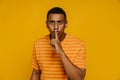 Afro man in striped orange shirt showing silence gesture while standing isolated