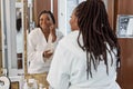 afro woman after shower stands in bathrobe look at mirrorapplying cream on facial skin feels happy Royalty Free Stock Photo