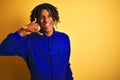 Afro worker man with dreadlocks wearing mechanic uniform over isolated yellow background smiling doing phone gesture with hand and Royalty Free Stock Photo