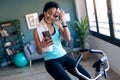 Afro young fitness girl using mobile phone while training on exercise bike at home