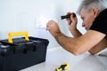 Aged electrician, repairman in uniform working, examining socket in the kitchen using flashlight. Repair service concept