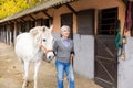 Aged farmer woman leading white horse outdoors along stables