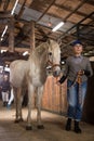 Aged female stable worker leading white horse by bridle in barn Royalty Free Stock Photo