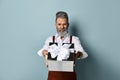 Aged male in white shirt, brown pants and suspenders, bracelet. He holding silver gift box, posing sideways on blue background Royalty Free Stock Photo