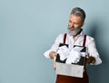 Aged male in white shirt, brown pants and suspenders, bracelet. He holding silver gift box, posing sideways on blue background Royalty Free Stock Photo
