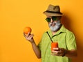 Aged man in hat, green shirt, sunglasses. Smiling, holding an orange, glass of fresh juice with tube, posing on orange background Royalty Free Stock Photo