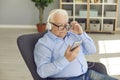 Aged man in shirt, glasses and headphones sitting in armchair and listening to music on smartphone