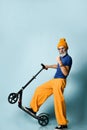 Aged man in t-shirt, sunglasses, orange pants, hat, gumshoes. Riding black scooter, showing fist, posing on blue background