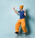 Aged man in t-shirt, sunglasses, orange pants, hat, gumshoes. Riding black skateboard, showing fists, posing on blue background