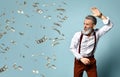 Aged man in white shirt, brown pants, suspenders, bracelet. He looks scared, swatting at hundred dollar bills, blue background