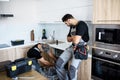 Aged repairman in uniform fixing dishwasher in the kitchen, while his young colleague standing near him and watching