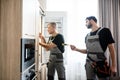 Aged repairman in uniform fixing refrigerator in the kitchen, while his colleague helping him, bringing screwdriver