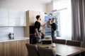 Aged repairman in uniform fixing refrigerator in the kitchen, while his colleague helping him, bringing screwdriver