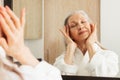 Aged woman with closed eyes touching her face with her fingers. Female in bathrobe doing face massage in bathroom Royalty Free Stock Photo