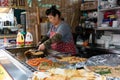 Aged woman cooking vegetable pancakes. Street food stall at a street market. She is using a grill and vegetable oil. Wearing apron