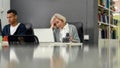 Aged woman, senior intern looking uncertain, confused at the screen while using laptop, sitting at desk, working in