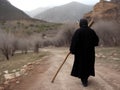An aged woman using a wooden stick to traverse an unpaved road lined by barren fields.. AI generation