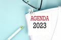 AGENDA 2023 text on a sticky on notebook with pen and glasses , blue background Royalty Free Stock Photo
