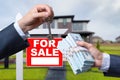 Agent Handing Over Keys as Buyer is Handing Over Cash for House with Home and For Sale Real Estate Sign Behind. Royalty Free Stock Photo