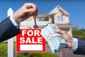 Agent Handing Over Keys as Buyer is Handing Over Cash for House with Home and For Sale Real Estate Sign Behind. Royalty Free Stock Photo