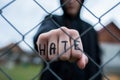 Aggressive teenage boy showing hes fist behind wired fence at the correctional institute, the word hate is written on hes hand