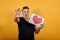 Aggressive young man keeping picture of heart, scream, showing palm at camera.