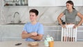 Frustrated offended guy sitting at kitchen table on background of swearing dissatisfied young wife. Family conflicts and