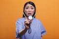 Agitated uneasy asian nurse using stethoscope to consult sick patient on orange background.