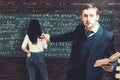 Agitated young professor pointing at green board with a lot of writing done by female student in white jumper and blue