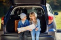 Agreeable boy and girl are looking at the road map while sitting in the auto`s trunk and discussing the move direction