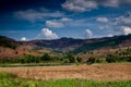 Agricultural field with a small hut and bald mountain range at the back against clear blue sky and white fluffy cumulus clouds.