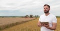 An agronomist stands with a tablet in a wheat field, a combine works in the background. A combine harvester harvests