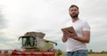 An agronomist stands with a tablet in a wheat field, a combine works in the background. A combine harvester harvests