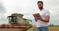 An agronomist stands with a tablet in a wheat field, a combine works in the background. A combine harvester harvests