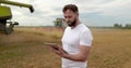 An agronomist stands with a tablet in a wheat field, a combine works in the background. A combine harvester harvests