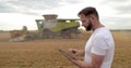 An agronomist stands with a tablet in a wheat field, a combine works in the background. A combine harvester harvests