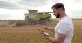 An agronomist stands with a tablet in a wheat field, a combine works in the background. A combine harvester harvests