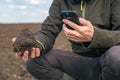 Agronomist is holding a clod of earth and smartphone, using the device to photograph the sample of the soil