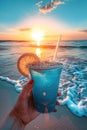 A hand holding a blue drink with a lemon slice against a sunset beach backdrop. Royalty Free Stock Photo