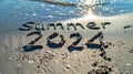 Summer 2024 written on a sandy shore, glistening water. Royalty Free Stock Photo