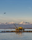 An airplane flies over a trebuchet fishing hut at sunset, against the snow-capped Alps, Marina di Pisa, Tuscany, Italy Royalty Free Stock Photo