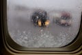 Airplane window with view partially blocked by falling snow. Blurred deicing trucks on the tarmac.