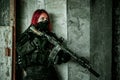 Airsoft red-hair woman in uniform with machine gun stand beside concrete wall. Soldier lurk inside broken building