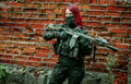 Airsoft red-hair woman in uniform with machine gun standing beside brick wall